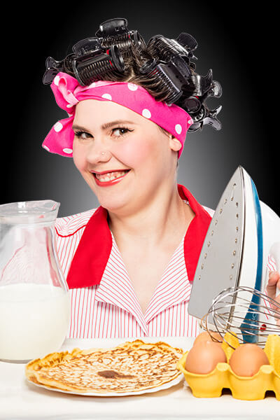 Key_Visual_Leistungen_Haushaltshilfe_400x600 junge Frau lächelt in Kamera, hat Lockenwickler im Haar und hält ein Bügeleisen und Eierkuchen vor sich