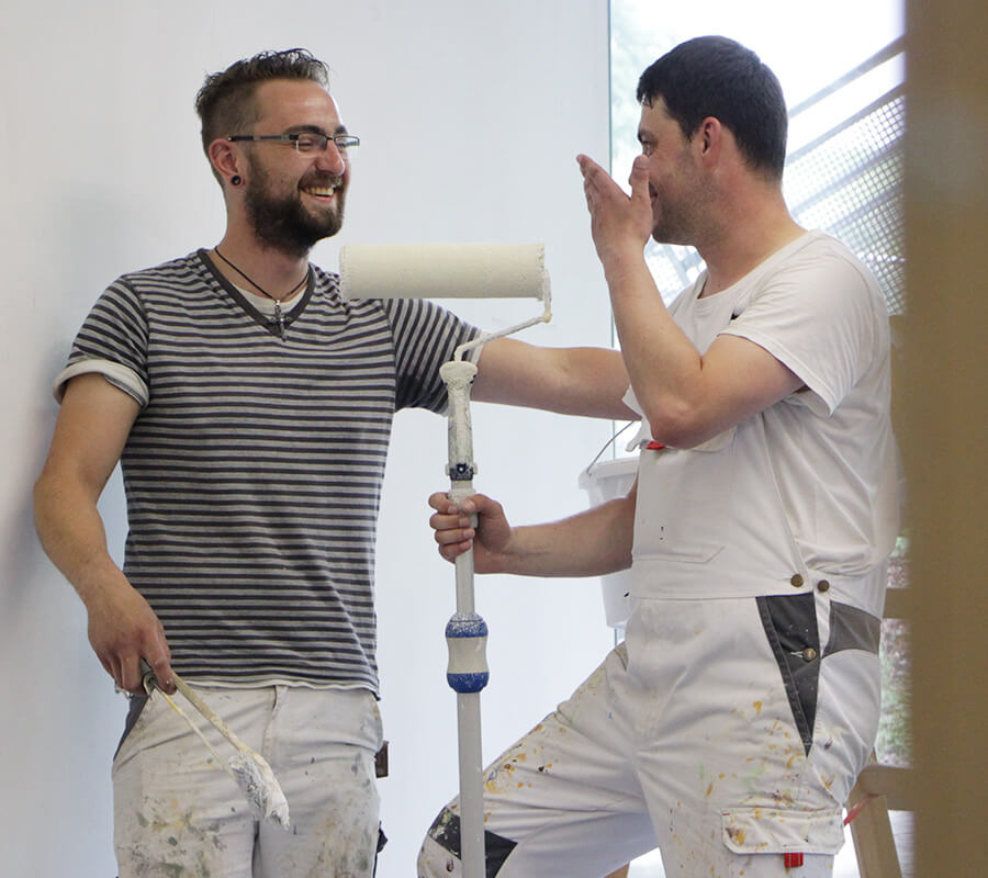 zwei junge Maler schauen lachen miteinander, der rechte hält einen Roller in der Hand